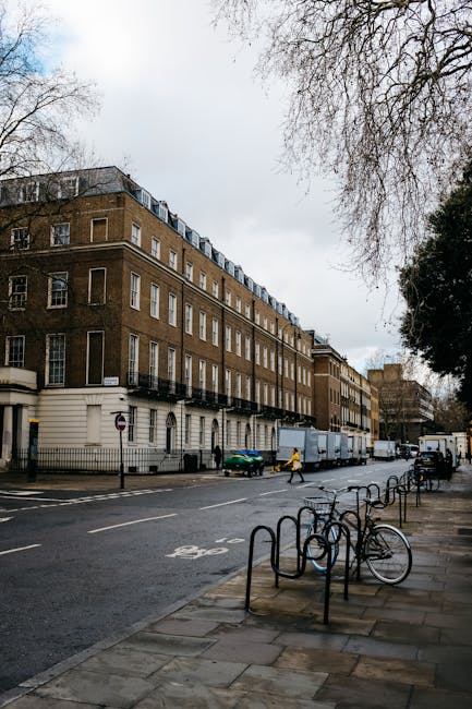 The image shows a quiet residential street with a row of elegant, multi-story Victorian-style buildings on the left, featuring large windows, decorative balcony railings, and a light-colored façade. 